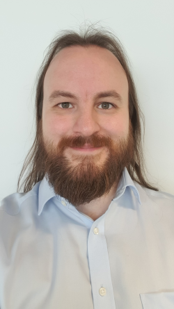 A picture of Matt Luckcuk taken in 2023. He is a white man with an oval face, long brown hair that comes over his shoulders, and a medium-length brown beard. He is smiling, slightly and looking straight into the camera lens. He is wearing a light blue shirt.